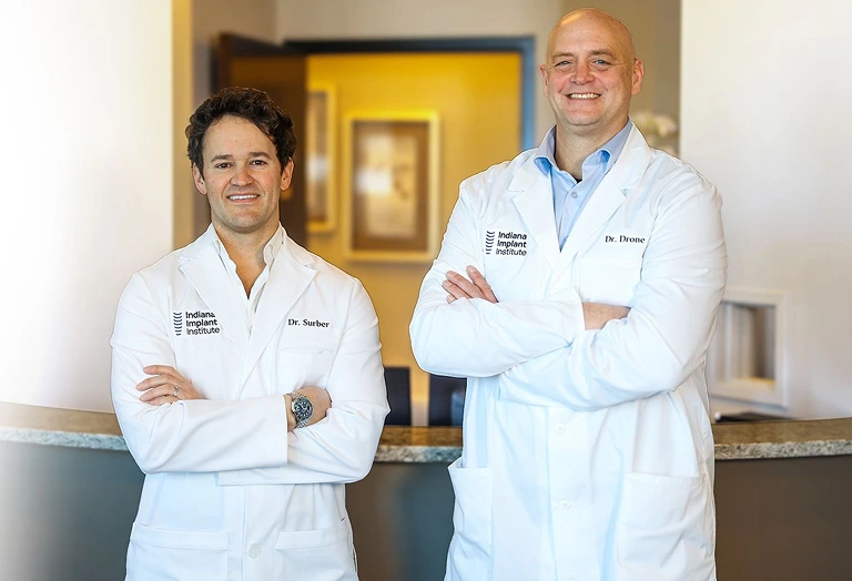 Dr. Surber and Dr. Drone, dental implant specialists at Indiana Implant Institute, smiling confidently in white lab coats.